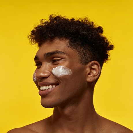 Cropped image of smiling black guy with cosmetic cream on his face looking away. Young brunette curly man. Facial skin care. Isolated on yellow background. Studio shoot. Copy space