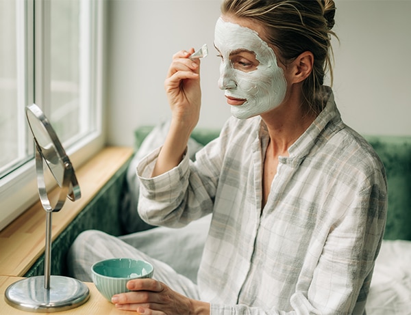 A woman in pajamas applies a clay mask to her face with a brush while looking in the mirror.