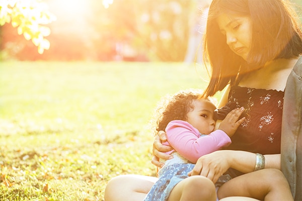 Hispanic mexican mother breastfeeding a dark-skinned mixed race toddler three year old daughter sitting on the green grass in nature - maternity concept. Little hispanic girl looking up. Copy space