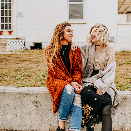 two women supporting each other smiling outside on wall on windy autumn day