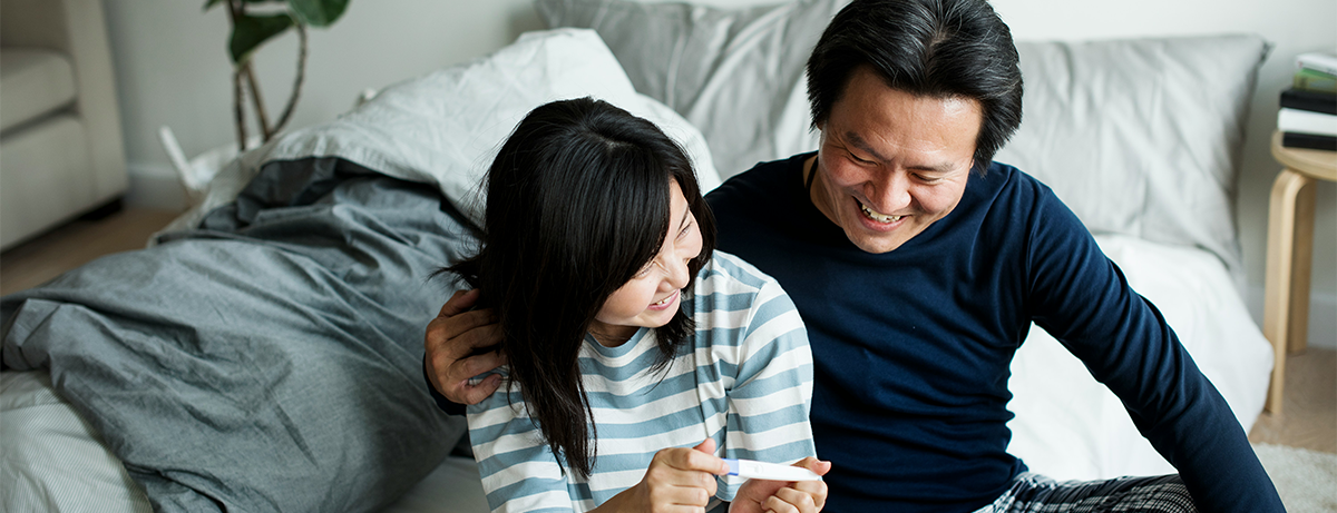 Asian male and female sitting on the bed with happy expressions. Female is holding a pregnancy test.