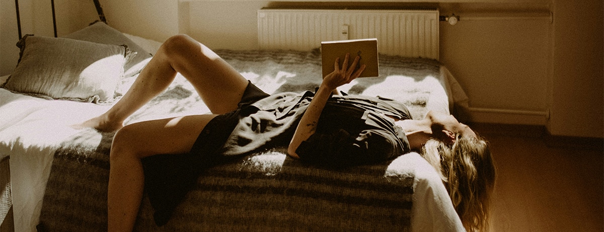 Female in black dress laying on bed and reading a book