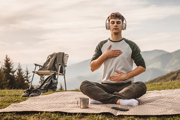 Young boy with headphones, meditate and sit down in front of tent on the mountain