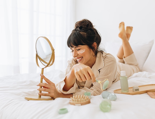 Close up of smiling young woman lying on bed and applying face cream. Woman doing beauty care face massage at home.