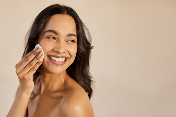 Beautiful young multiethnic woman with bare shoulder removing make up with cotton pad on cheek isolated against beige background. Smiling hispanic girl applying dry powder using cosmetic cushion.