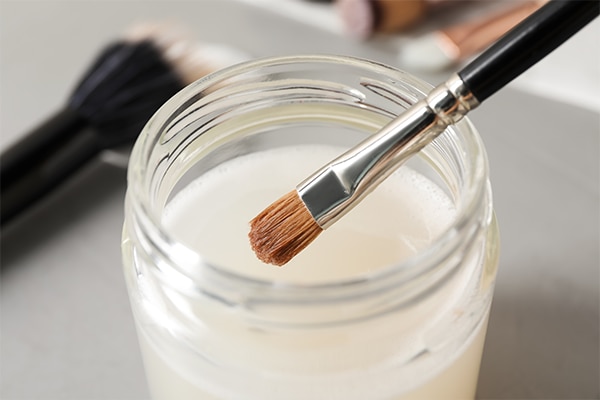 Cleaning makeup brush in jar with special liquid on grey table, closeup