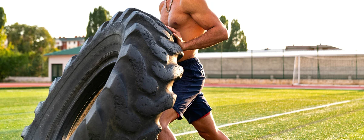 A man is lifting a tire on a field. He is wearing a necklace and has a muscular build