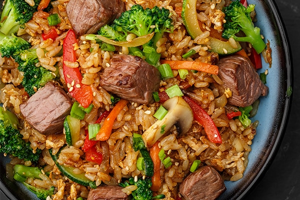 Asian rice with teriyaki beef and vegetables: zucchini, broccoli, pepper, mushrooms, carrot, onion and sesame seeds. Dish isolated in a blue bowl, close-up on a black marble background. Asian cuisine.
