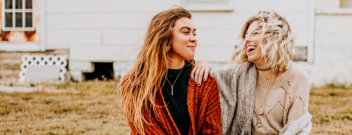 two women supporting each other smiling outside on wall on windy autumn day