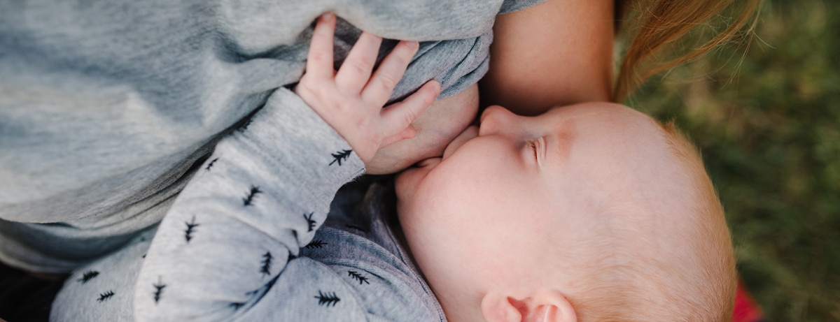 Breastfeeding baby. Mother breast feeding newborn boy sitting on grass in summer park. Concept of lactation infant, postpartum period, natural motherhood. Mom hug son on nature. Babywearing. Closeup.
