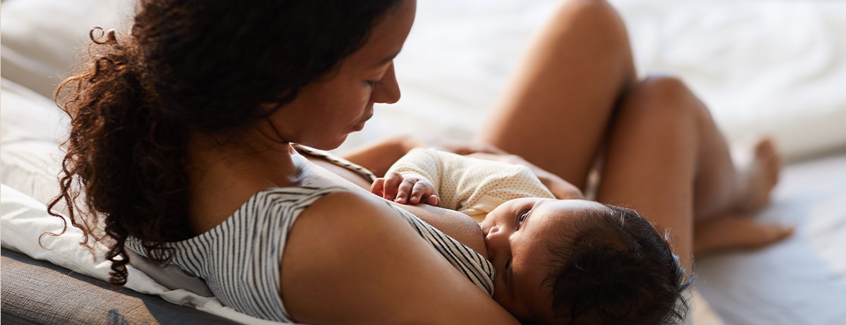 Peaceful loving young African mother sitting on bed and leaning on headboard while feeding baby with breast