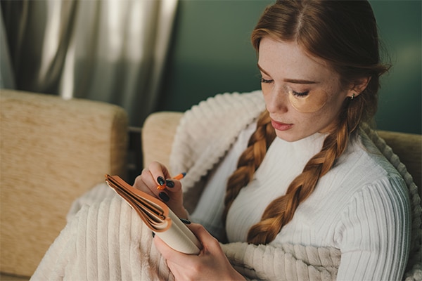 Young minded woman wrapped in plaid writing down memories in notebook sitting on sofa at home spending free spare time in living room. Autumn season mood.