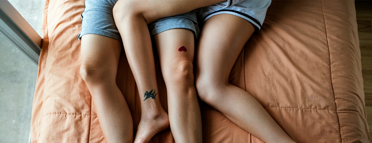 Female couples legs in bed. Both have leg tattoos on a brown duvet