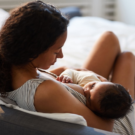 Peaceful loving young African mother sitting on bed and leaning on headboard while feeding baby with breast