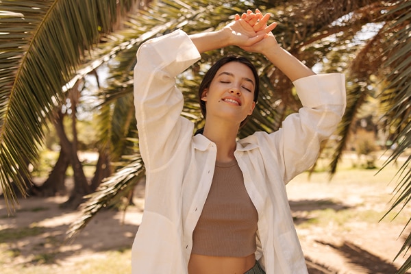 Relaxed young caucasian girl basking in sun with eyes closed, holding hands above head standing in nature. Brunette wears grey top, white shirt. Summer vacation concept.