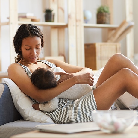 Serious calm careful young black mom sitting on sofa bed and breastfeeding baby while holding son in arms