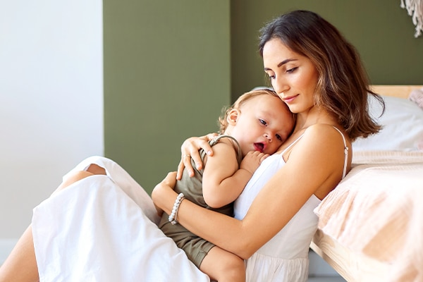Young beautiful woman mother hugging embracing cute baby boy while sitting together on floor in bedroom near bassinet. Mom cuddling her little son infant and expressing love and care.