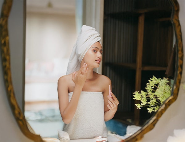 Indian young adult gen z beautiful woman wrapped towel on head and body look bathroom mirror hold blade razor tool shave her face remove facial hair do beauty skincare routine day indoor modern home