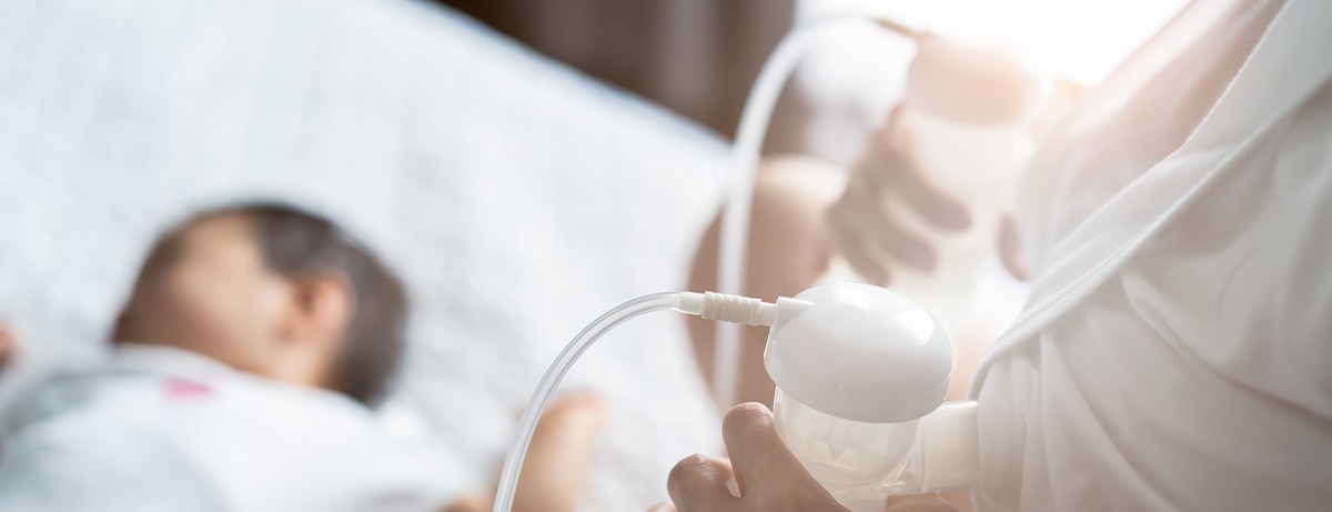 Asian pretty mother pumping breast milk on the bed in bedroom. The baby lying and sleeping on the bed beside her. Mother holding the milk and looking the child with love and smiling. Pump milk concept