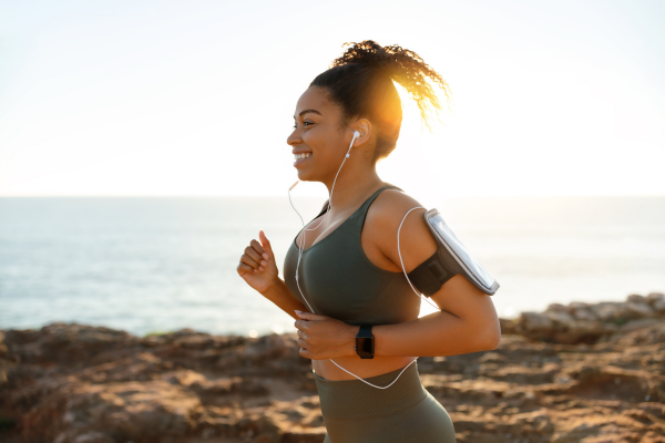 Happy young african american female in sports wear, headphones with smartphone running in morning on rocks near ocean alone, outdoor. Health care, training, active lifestyle and sports with music